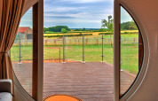 Zicht vanuit een pod door een ronde deur op een terras en groene velden in het landelijke landschap.