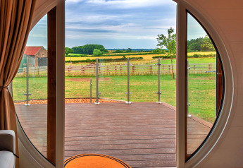 Zicht vanuit een pod door een ronde deur op een terras en groene velden in het landelijke landschap.