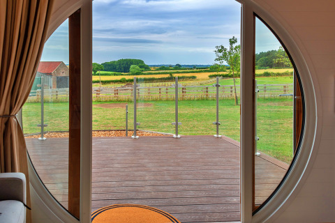 View from inside a pod through a circular door, overlooking a deck and lush green countryside beyond.