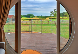 View from inside a pod through a circular door, overlooking a deck and lush green countryside beyond.