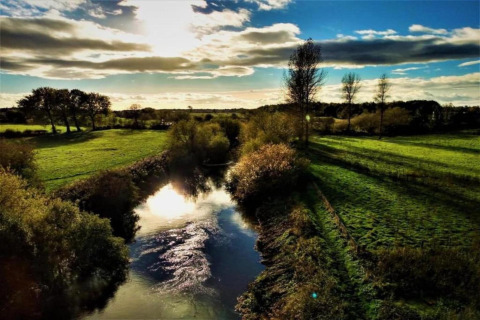 Photo de paysage d’un étang avec reflet du soleil, champs verts et arbres sous un ciel partiellement nuageux.