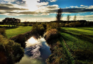 Photo de paysage d’un étang avec reflet du soleil, champs verts et arbres sous un ciel partiellement nuageux.