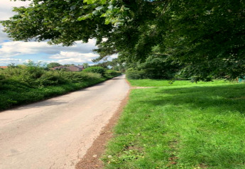 Landweg omringd door fris groen gras en bomen, met zicht op een dorp onder een deels bewolkte hemel.
