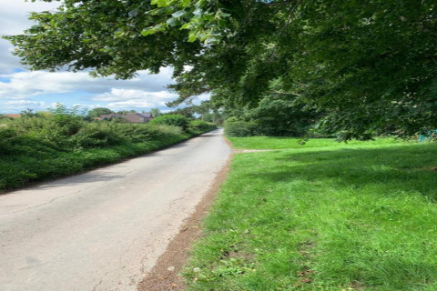 Route de campagne bordée d'herbe verte et d'arbres, menant vers un village sous un ciel partiellement nuageux.