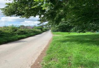 Route de campagne bordée d'herbe verte et d'arbres, menant vers un village sous un ciel partiellement nuageux.