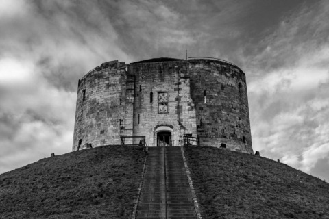 Photo en noir et blanc d’une vieille tour circulaire en pierre sur une colline avec escalier, ciel dramatique.