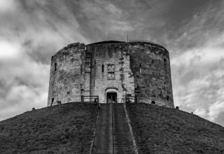 Photo en noir et blanc d’une vieille tour circulaire en pierre sur une colline avec escalier, ciel dramatique.