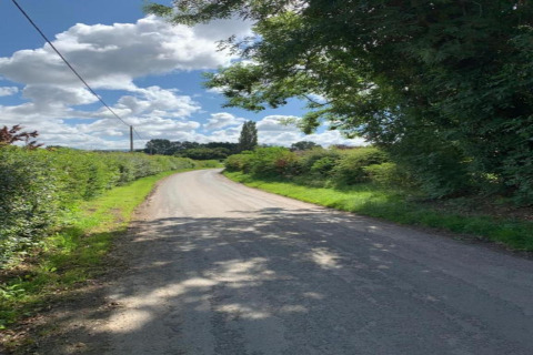 Une route de campagne sinueuse bordée de haies et d’arbres sous un ciel bleu partiellement nuageux.