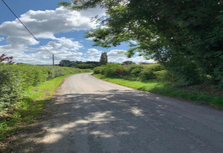 Une route de campagne sinueuse bordée de haies et d’arbres sous un ciel bleu partiellement nuageux.