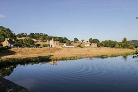 Paysage rural avec un étang au premier plan, des ruines et des champs sous un ciel bleu clair.