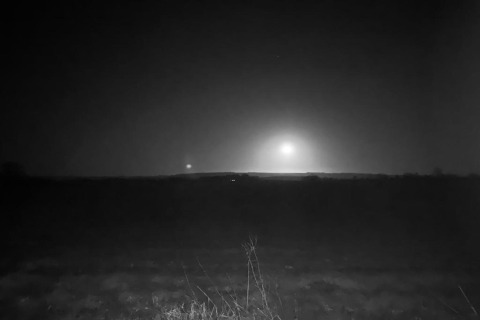 Photo nocturne en noir et blanc d'un étang avec de l'herbe au premier plan et une lumière brillante à l'horizon.
