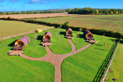 Vier glampingpods op een grasveld met paden, omringd door open landschap en landelijke natuur.