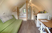 Intérieur d’un pod moderne avec canapé vert, lit double, petite cuisine, coin repas et salle de bain.