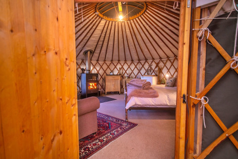 Vista interna della Bramble Yurt nel Regno Unito con letto confortevole, stufa a legna e atmosfera accogliente.