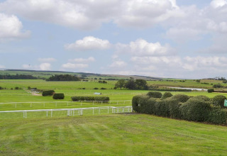 Foto van een groot groen veld met heggen en afrasteringen onder een bewolkte lucht, mogelijk een renbaan.