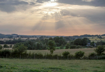 Landschapsfoto genomen aan een pod, met groene velden, bomen en zonnestralen doorheen de wolken.