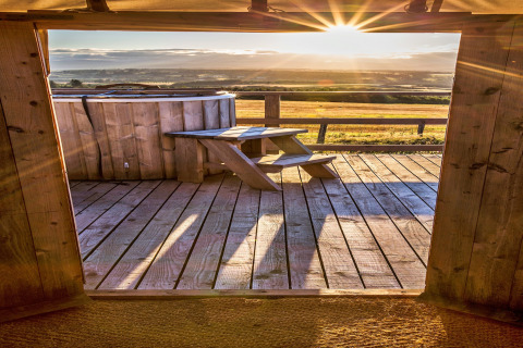 View from Bramble Yurt in the UK, showing a wooden deck, hot tub, and sunset over open countryside.