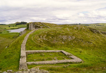 Photo d’un ancien mur de pierre près d’un étang dans un paysage vallonné verdoyant sous un ciel nuageux.