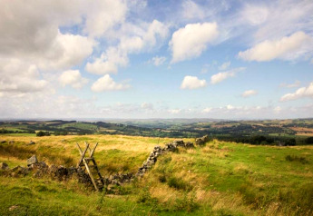 Zicht op het groene Engelse landschap met een stenen muur en houten overstap onder wolkenlucht.