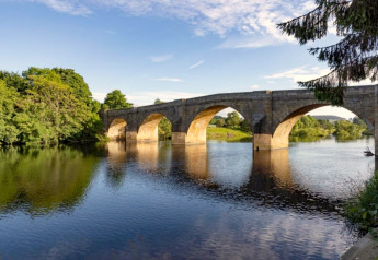 Pont de pierre avec arches traversant une rivière paisible, entouré de verdure sous un ciel bleu clair.