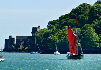 Barca a vela con vela rossa e yacht sull’acqua vicino a un castello tra alberi verdi rigogliosi.