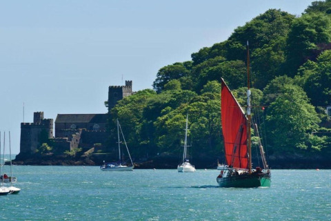 Voilier à voile rouge et yachts sur l'eau près d'un château entouré d'arbres verts luxuriants.