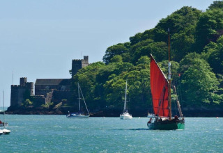 Voilier à voile rouge et yachts sur l'eau près d'un château entouré d'arbres verts luxuriants.