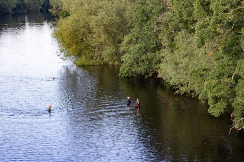 Mensen zwemmen en staan in een brede rivier naast groene bomen op een rustige dag in de natuur.