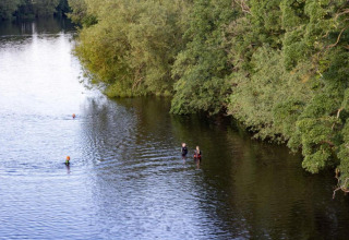 Mensen zwemmen en staan in een brede rivier naast groene bomen op een rustige dag in de natuur.