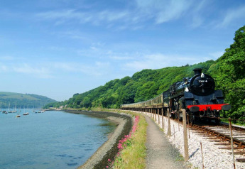 Un treno a vapore viaggia lungo il fiume vicino a Oak Tree Lodge nel Regno Unito tra colline verdi.