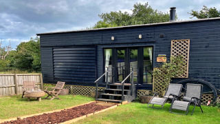 Mobilhome Oak Tree Lodge med sort facade, havemøbler og liggestole i grøn have under overskyet himmel.