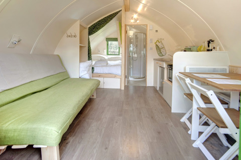 Interieur van een moderne, knusse pod met een groene bank, eettafel, kleine keuken en slaapgedeelte.