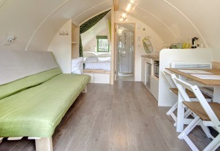 Interieur van een moderne, knusse pod met een groene bank, eettafel, kleine keuken en slaapgedeelte.