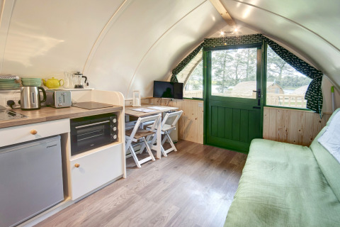Knus pod-interieur met kitchenette, kleine eettafel, bank en groene deur met uitzicht op buitenleven.