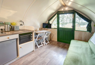 Knus pod-interieur met kitchenette, kleine eettafel, bank en groene deur met uitzicht op buitenleven.