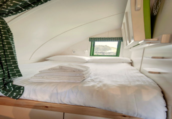 Binnenzicht van een knusse slaapruimte in een pod met wit beddengoed, groene gordijnen en een venster.