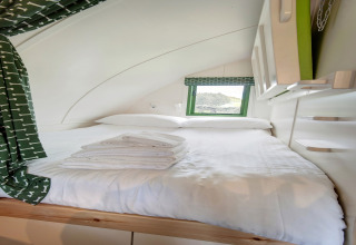 Interieur van een gezellige slaapruimte in een pod met wit beddengoed, groene gordijnen en een klein raam.