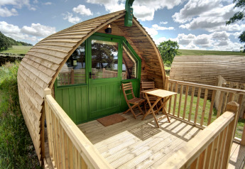 Houten glamping pod met groene deur en terras met tuinmeubelen in het zonnige Britse landschap.