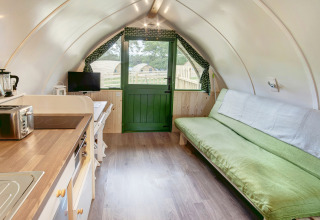 Modern pod-interieur met kleine keuken, groene slaapbank, eethoek, tv en deur met uitzicht naar buiten.
