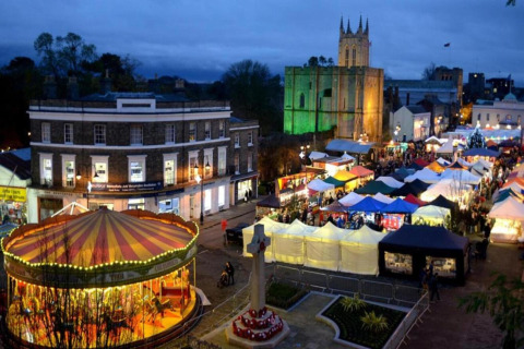 Avondbeeld van een feestelijke markt met een verlichte draaimolen, kleurrijke kraampjes en een oude kerk.