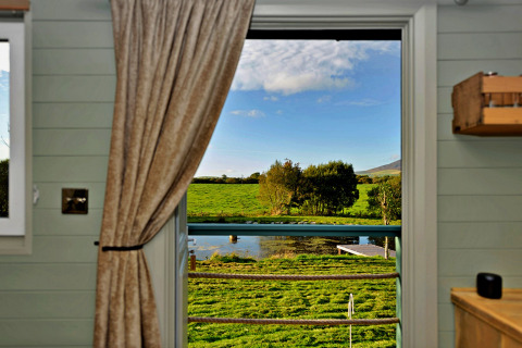 Uitzicht vanuit de 'Joan' Shepherds Hut over groene velden en een vijver door een open raam in het Verenigd Koninkrijk.