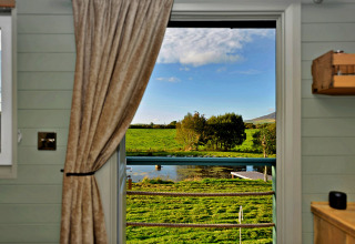 Uitzicht vanuit de 'Joan' Shepherds Hut over groene velden en een vijver door een open raam in het Verenigd Koninkrijk.