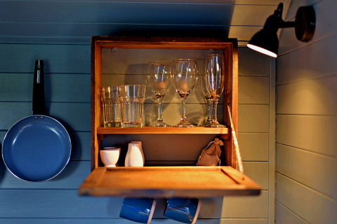 Interno di casa mobile con mensola in legno, bicchieri, tazze e padella blu appesa alla parete.