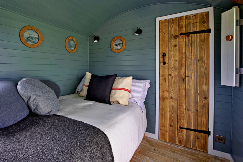 Interno di una roulotte accogliente con porta di legno, pareti verdi, oblò rotondi e letto con cuscini.