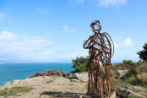 Scultura di metallo su una scogliera con vista mare, foto scattata in una struttura glamping con cielo sereno.