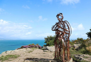 Metal sculpture on a rocky cliff overlooking the sea, taken at a glamping site under a clear blue sky.