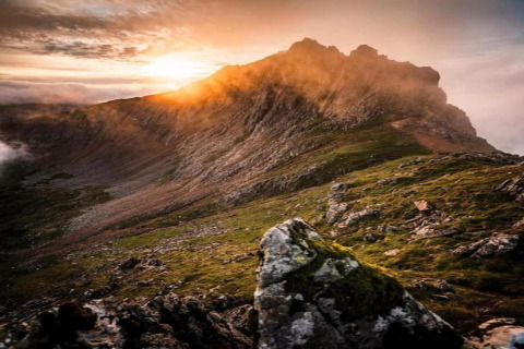 Vetta di montagna illuminata dal tramonto, luce calda su rocce e pendii erbosi e suggestivi.