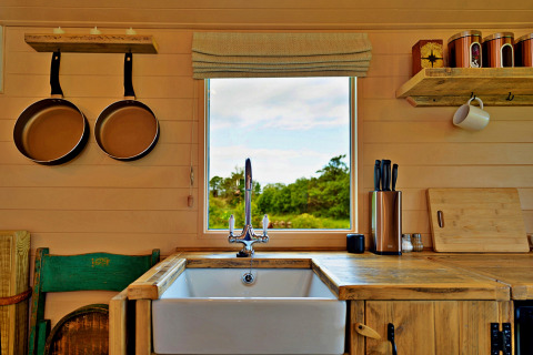 Tiny house kitchen with wooden counter, farmhouse sink, window view, frying pans, and utensils.