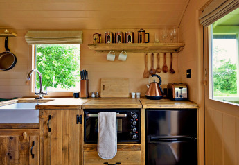 Cozy tiny wooden kitchen in the 'Marared' Gypsy Hut, UK, featuring rustic decor and leafy outdoor view.
