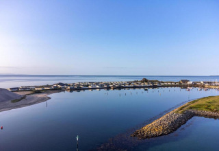 Panoramic view of a glamping site by the water, featuring cabins, calm waves, and a clear blue sky.
